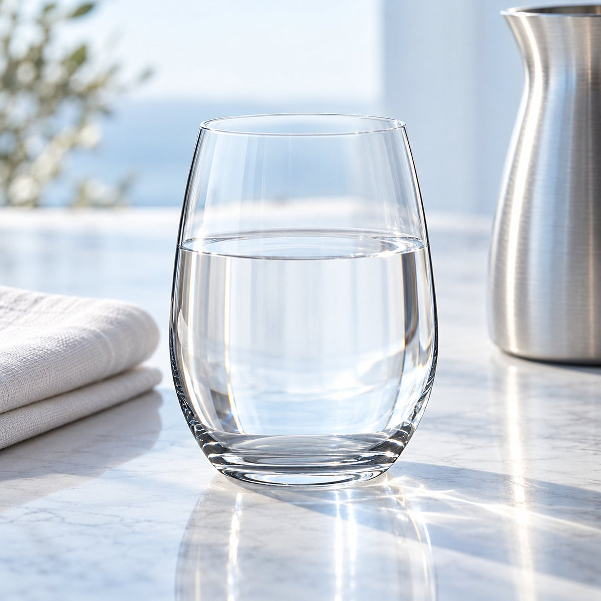 Verre à eau Nude Pure rempli d'eau à côté d'une carafe métallique et d'une serviette