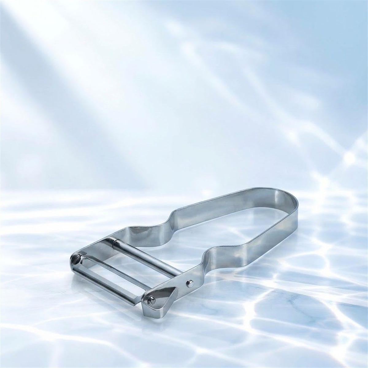 Économe inox à lame plate posé en diagonale sur un marbre blanc avec reflets d'eau, fond bleu clair