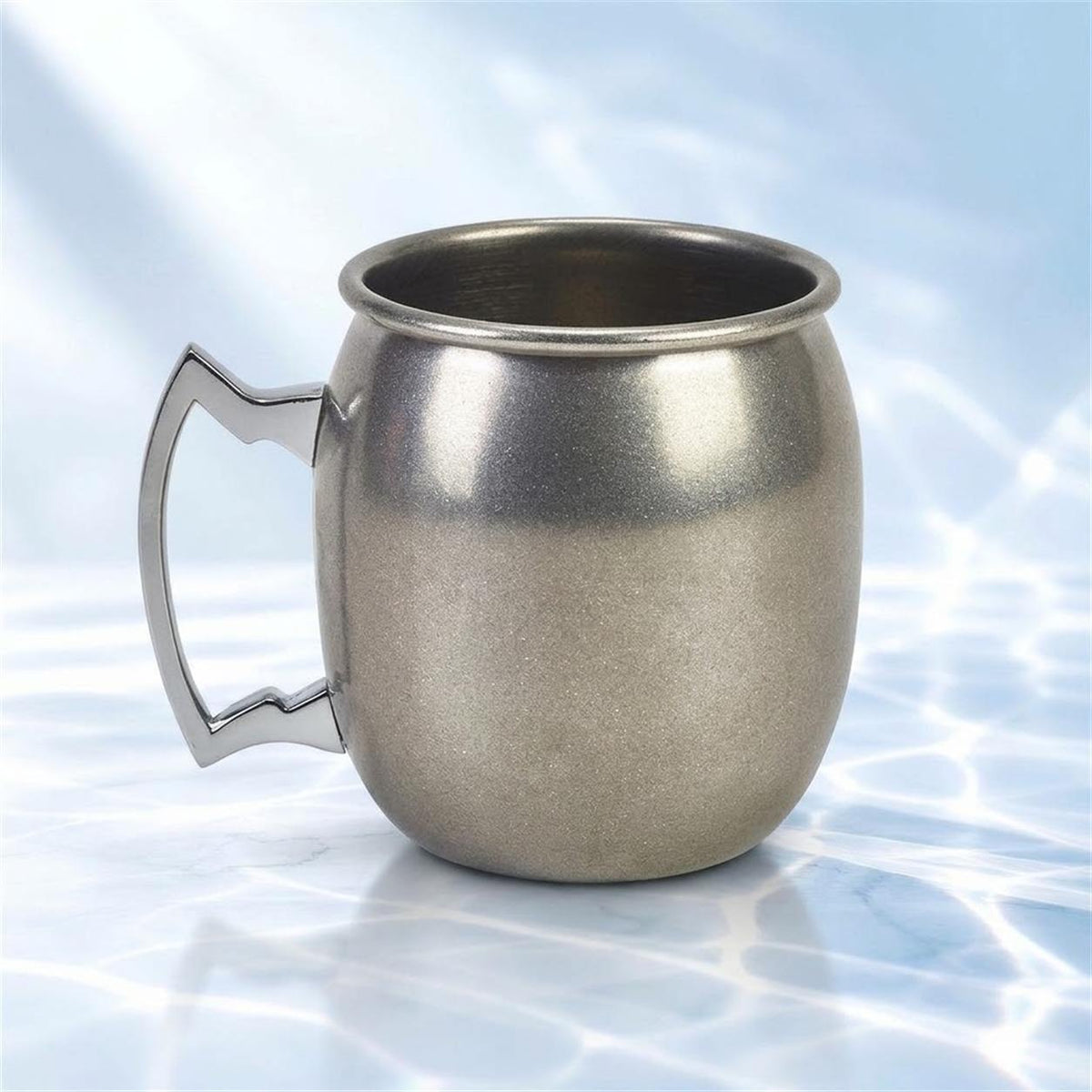 Mug Moscow mule en forme de tonneau, argent vieilli et poignée inox, sur un marbre blanc avec reflets d'eau, fond bleu clair