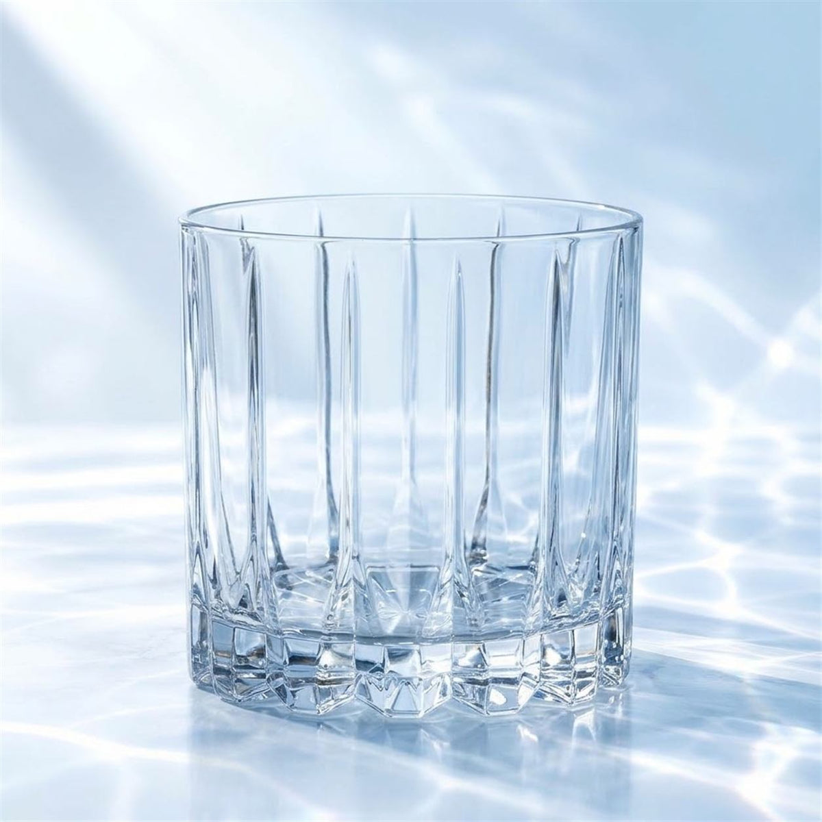 Verre short drink Riedel Drink Specific à nervures taillées, vide, debout sur un marbre blanc avec reflets d'eau, fond bleu clair