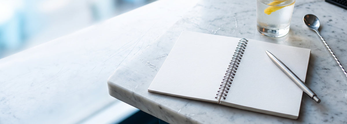 Blog BWA, actualités et conseils du bar : carnet ouvert, stylo, verre de cocktail et cuillère de bar sur un comptoir en marbre